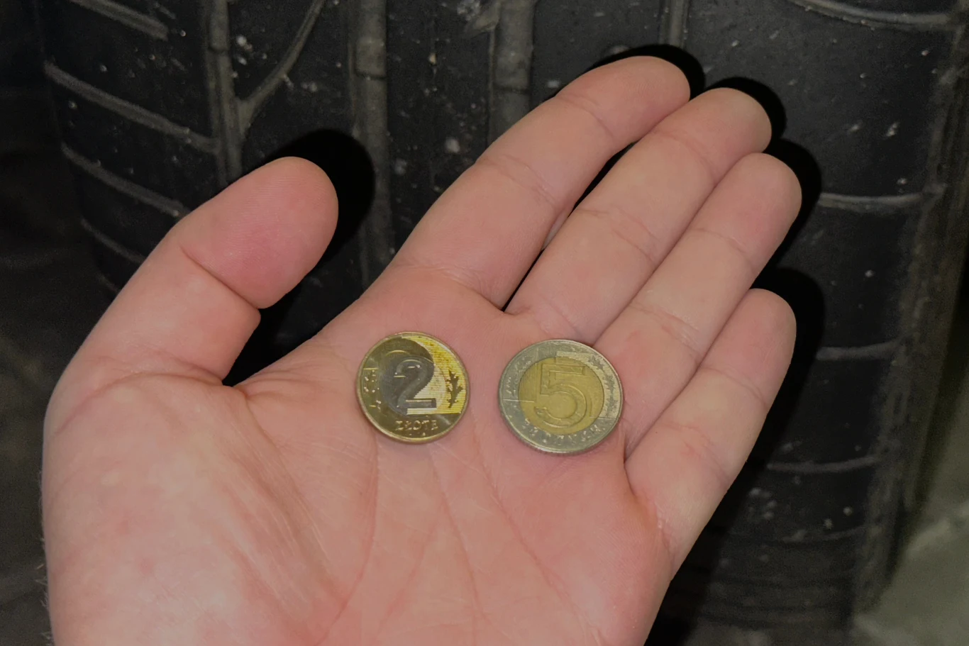 Dłoń trzymająca dwie monety o nominale 2 i 5 złotych na tle opony samochodowej.