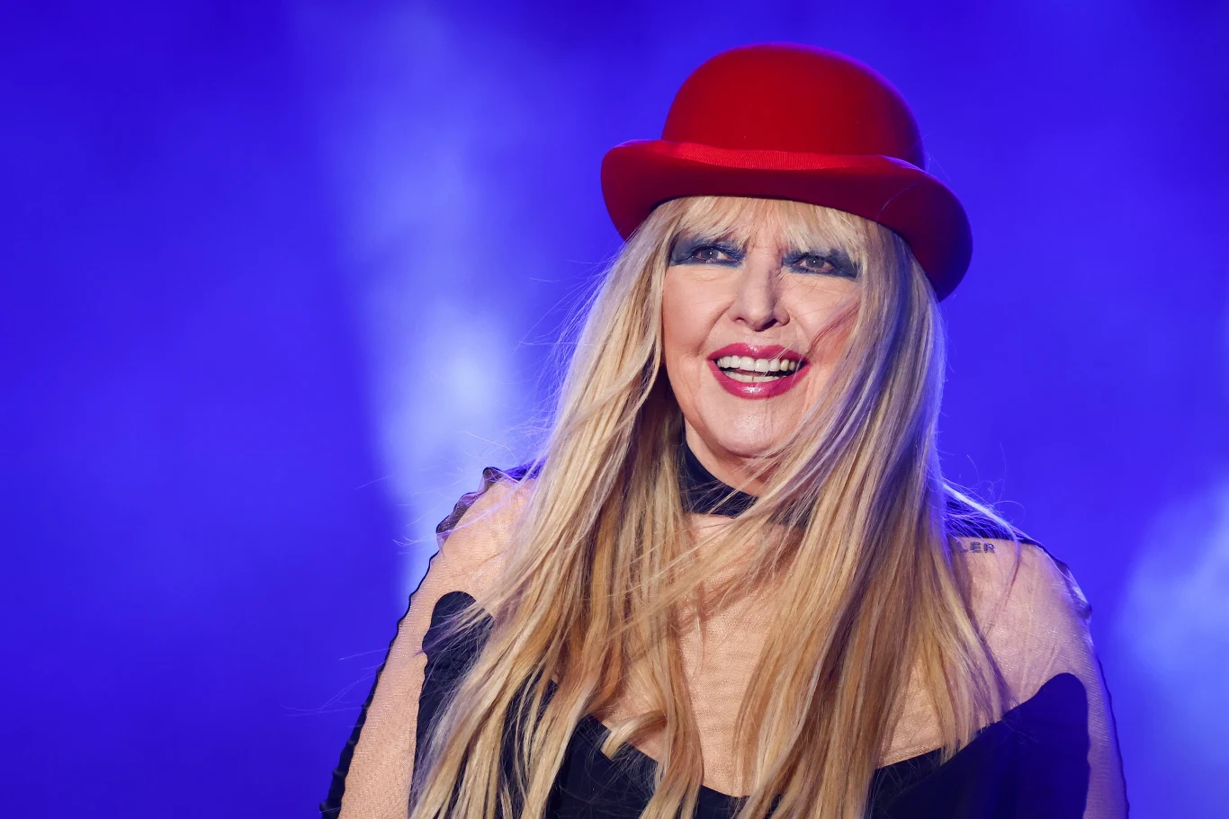 Maryla Rodowicz nie była pierwszą wykonawczynią "Kolorowych jarmarków"! Kobieta w średnim wieku z długimi blond włosami, uśmiechnięta, ubrana w ciemny strój i czerwoną melonową czapkę na głowie, znajduje się na tle intensywnie niebieskich świateł scenicznych.