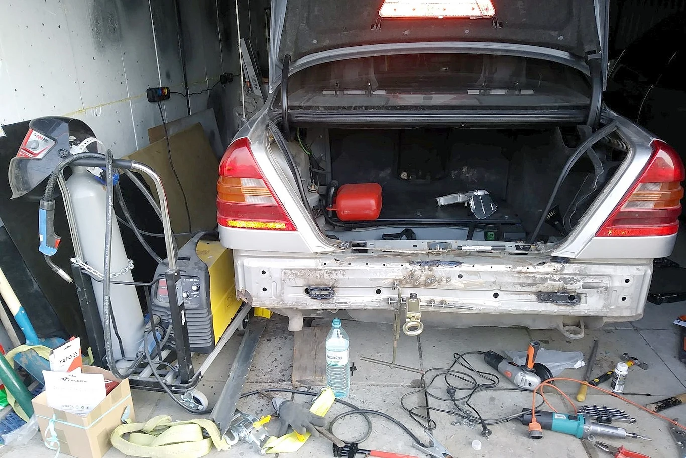 Szybka naprawa pokolizyjna w "warunkach domowych" / archiwum własne autora Otwarty bagażnik srebrnego samochodu osobowego w garażu, wokół rozrzucone narzędzia elektryczne, przewody, spawarka, butelka z wodą i skrzynka z częściami, widoczna czerwona kanister w bagażniku.