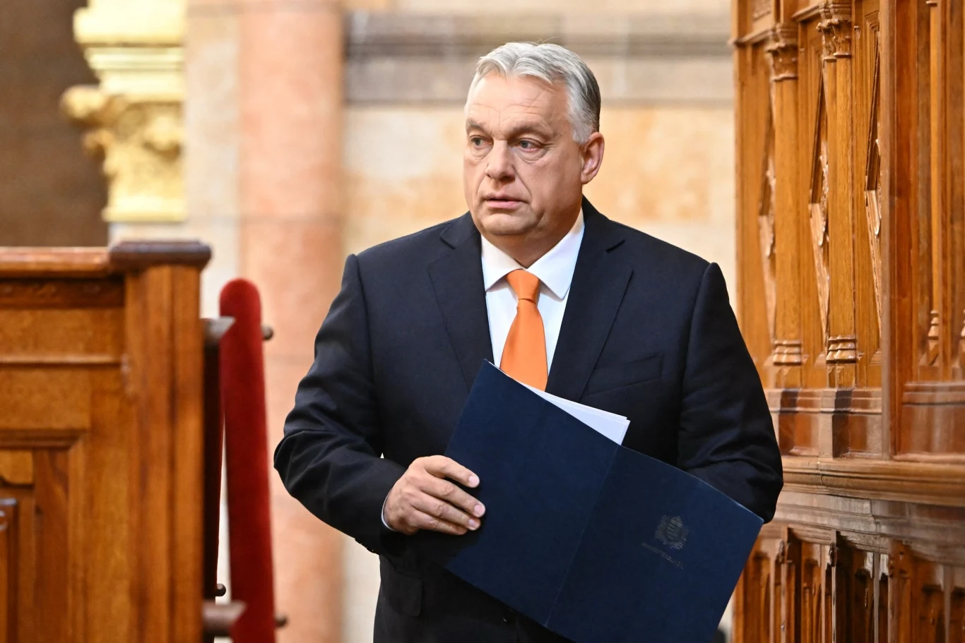 Premier Viktor Orban stwierdził, że wojna w Ukrainie kosztowała Węgry 20 mld euro Mężczyzna w garniturze z pomarańczowym krawatem trzymający teczkę w rękach, stojący wewnątrz budynku o drewnianych elementach architektonicznych.