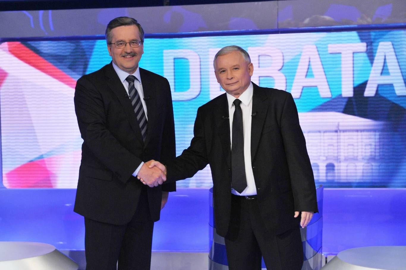 Dwóch mężczyzn ubranych w garnitury podaje sobie ręce na tle napisu 'DEBATA', w kadrze widoczne fragmenty flagi w tle oraz podświetlona scena.