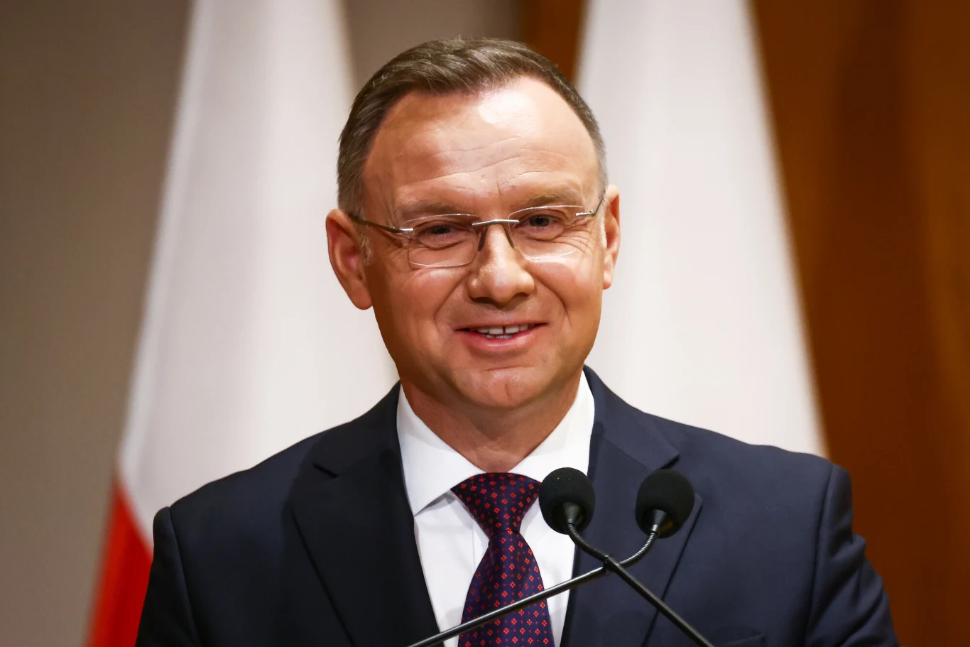 Polacy ocenili pracę Andrzeja Dudy Mężczyzna w garniturze i krawacie przemawia do mikrofonów, w tle widoczne rozmyte biało-czerwone flagi.