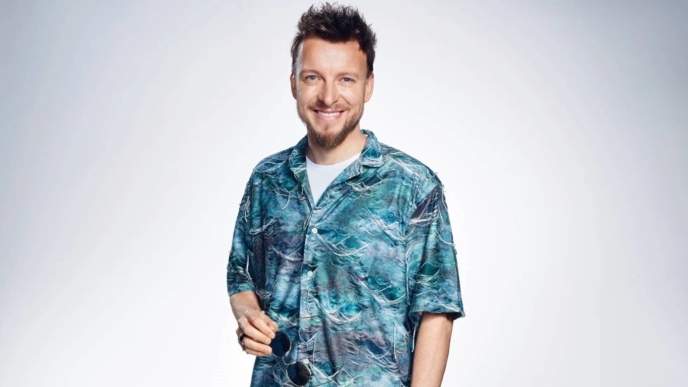 Piotr Kupicha Uśmiechnięty mężczyzna z brodą w hawajskiej koszuli z niebiesko-zielonym wzorem, trzyma w dłoni okulary przeciwsłoneczne; jasne, neutralne tło.