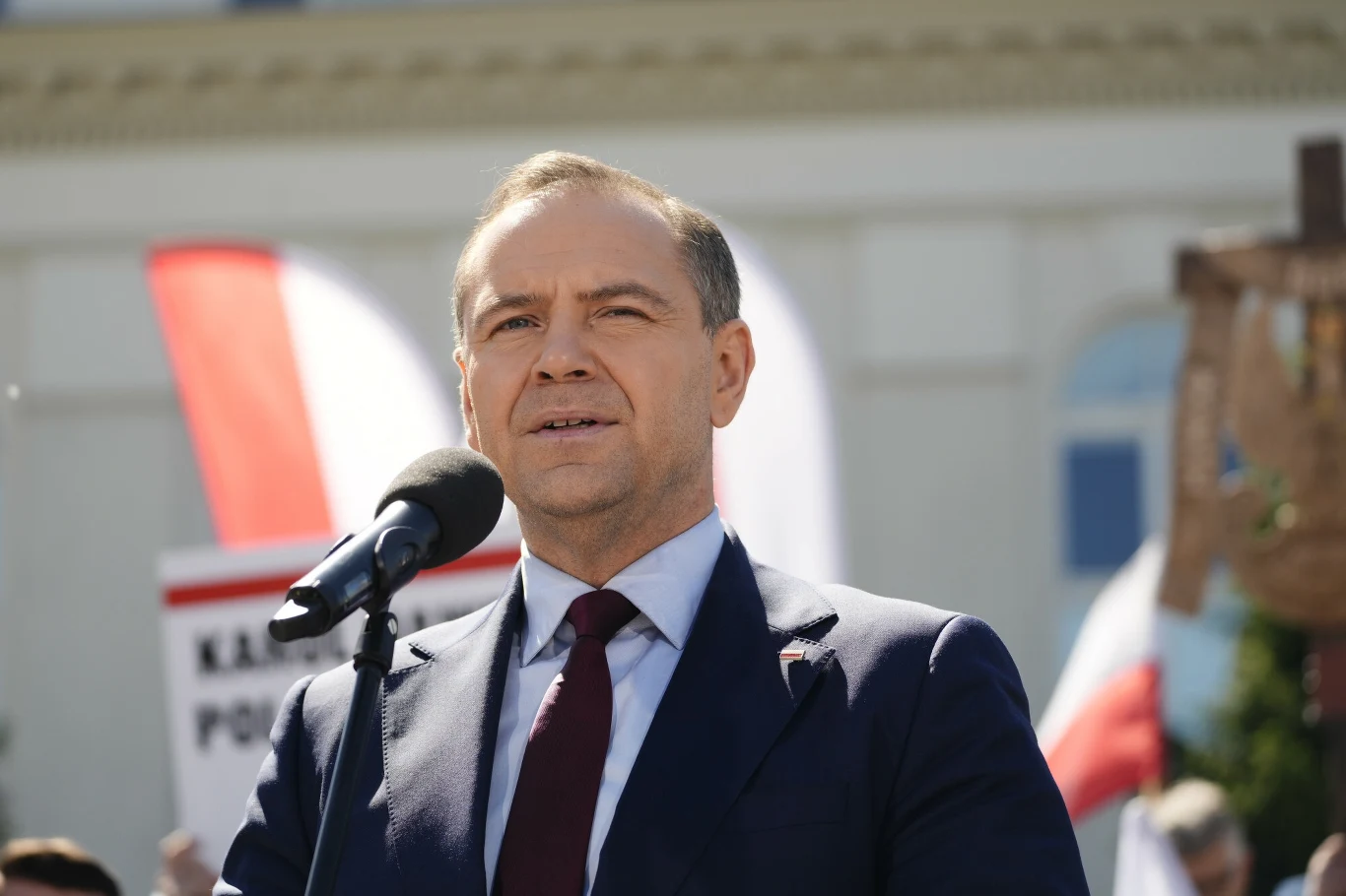 Wybory prezydenckie 2025. Debata. Padło pytanie o ustawki kibicowskie Mężczyzna w garniturze przemawia do mikrofonu na tle biało-czerwonych flag oraz tablicy z napisem, w słoneczny dzień na zewnątrz budynku.