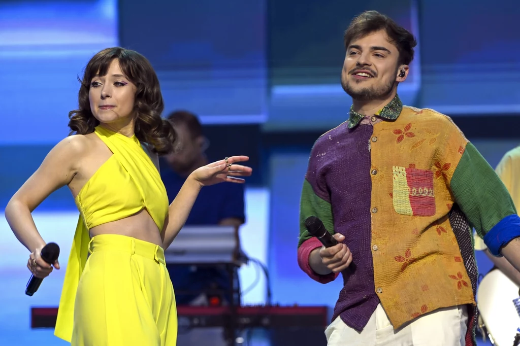 Wiktor Dyduła i Katarzyna Sienkiewicz Dwoje młodych wokalistów występujących na scenie z mikrofonami w rękach, kobieta ubrana w jasnożółtą sukienkę, mężczyzna w kolorowej koszuli, w tle widoczny sprzęt muzyczny i klawiatura, scena oświetlona niebiesko-fioletowym światłem.