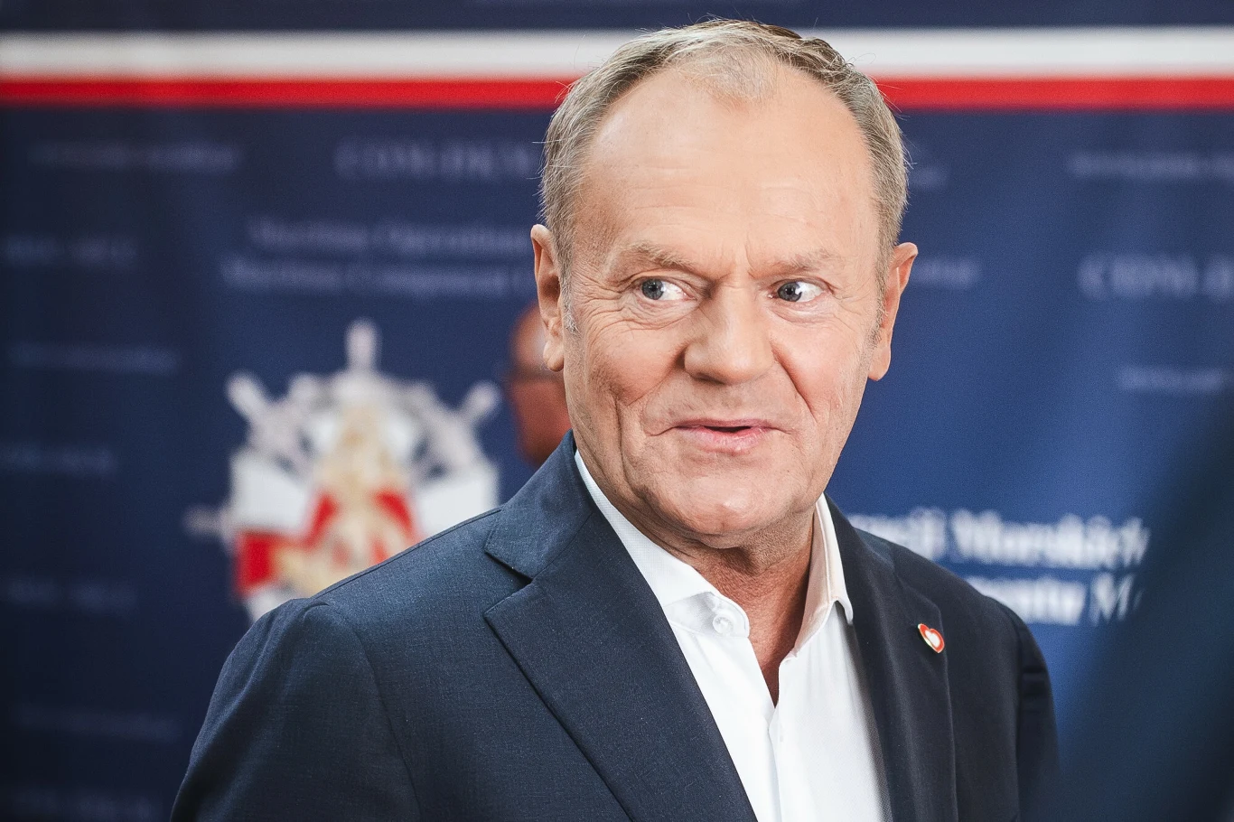 Donald Tusk zabrał głos po debacie Mężczyzna w średnim wieku w garniturze i białej koszuli, na tle niebieskiej tablicy z nieczytelnymi napisami oraz herbem, spogląda na bok z lekkim uśmiechem.