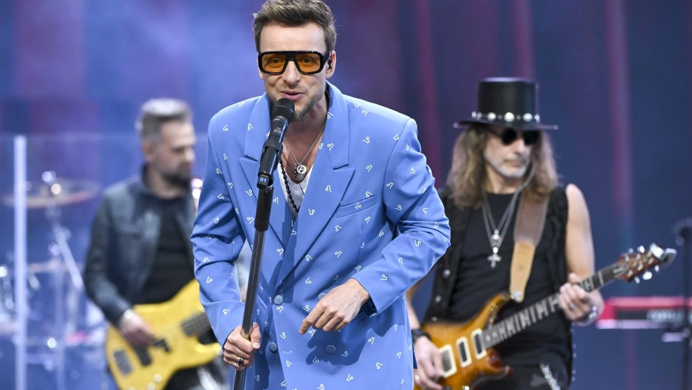 Mężczyzna w jasnoniebieskim garniturze i dużych okularach śpiewa do mikrofonu, za nim muzycy grający na gitarze basowej i gitarze elektrycznej, scena koncertowa, reflektory i dym w tle.