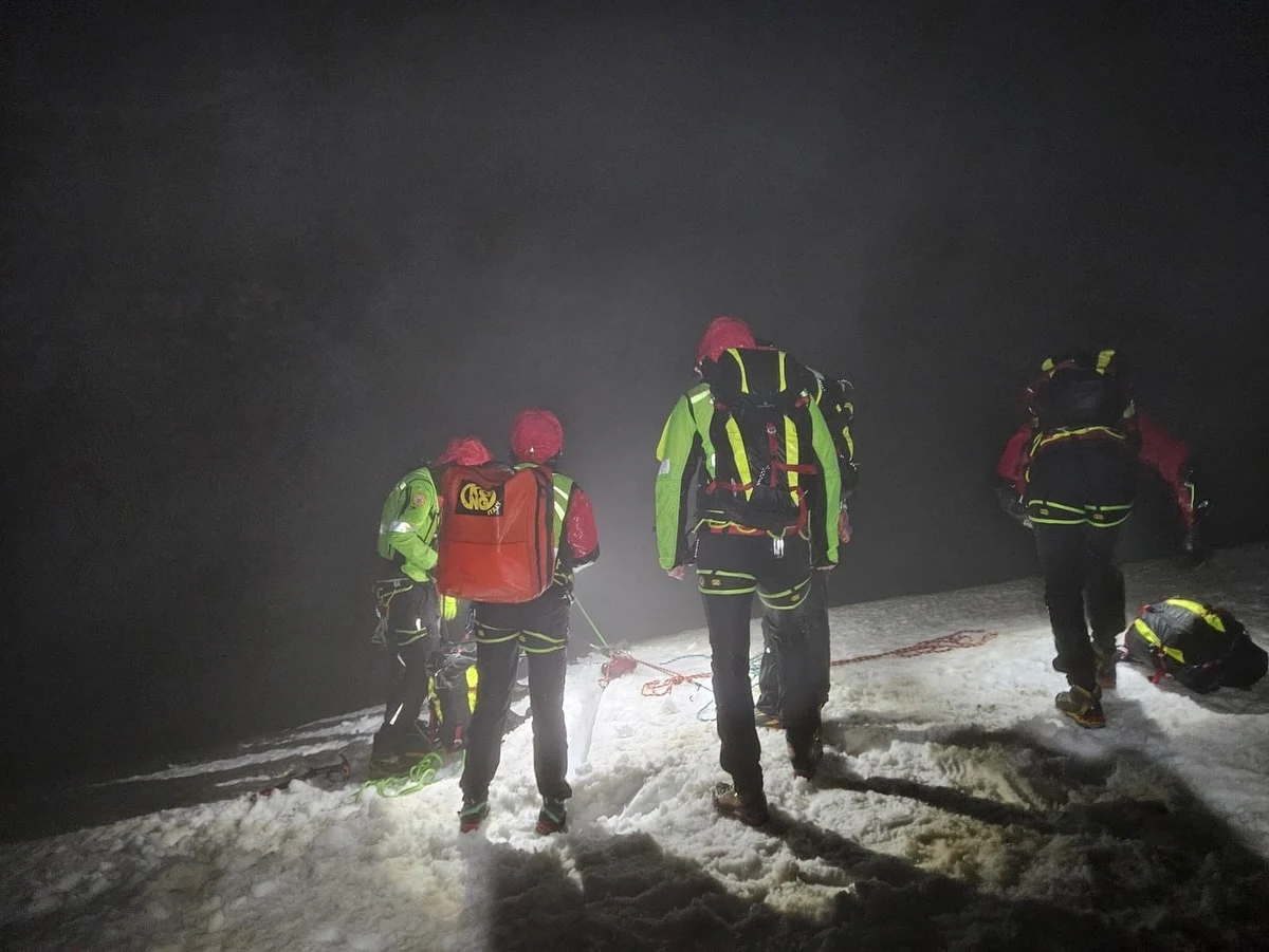 Włochy. Trudna akcja ratunkowa w górach. Udało się dotrzeć do polskiego paralotniarza Grupa ratowników górskich w specjalistycznych ubraniach oraz z latarkami i sprzętem ratunkowym prowadzi akcję ratunkową na zaśnieżonym zboczu podczas nocy, używając lin asekuracyjnych.