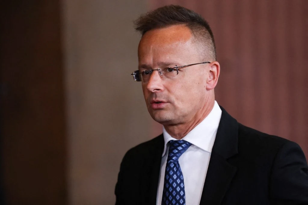 Szef węgierskiej dyplomacji Peter Szijjarto Mężczyzna w garniturze i krawacie o wzorze geometrycznym, krótkie włosy, okulary, poważny wyraz twarzy, jednolite tło w odcieniach brązu.