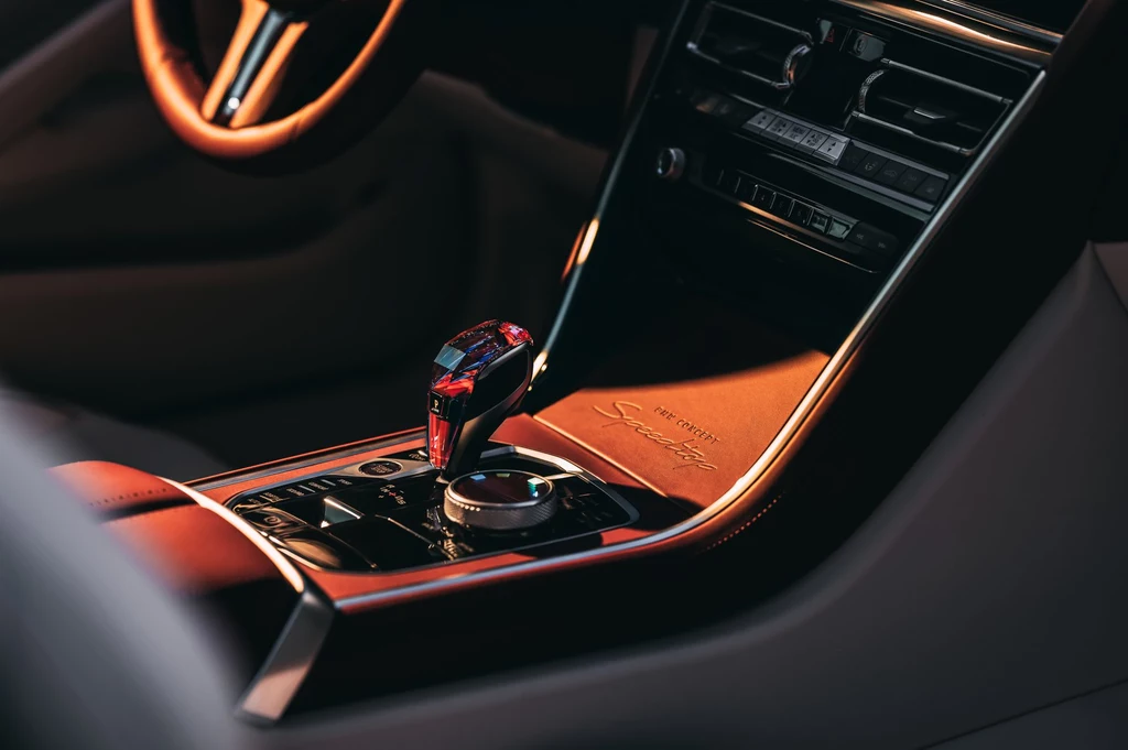 Centralna konsola z kryształowym wykończeniem, emblematem modelu i klasycznymi przełącznikami BMW – rzemiosło w czystej postaci. Luksusowa konsola środkowa samochodu z elegancką dźwignią zmiany biegów, skórzanym wykończeniem i detalami w ciemnych, stonowanych kolorach oświetlonych naturalnym światłem.