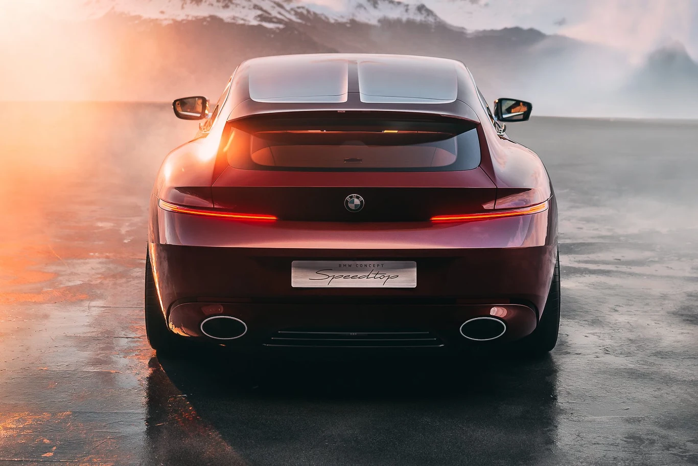 Czerwony sportowy samochód widziany od tyłu, z charakterystycznymi poziomymi światłami i dwoma szerokimi rurami wydechowymi, stoi na wilgotnej drodze, w tle rozmyte góry i atmosfera o zachodzie słońca dodająca dramatyzmu.