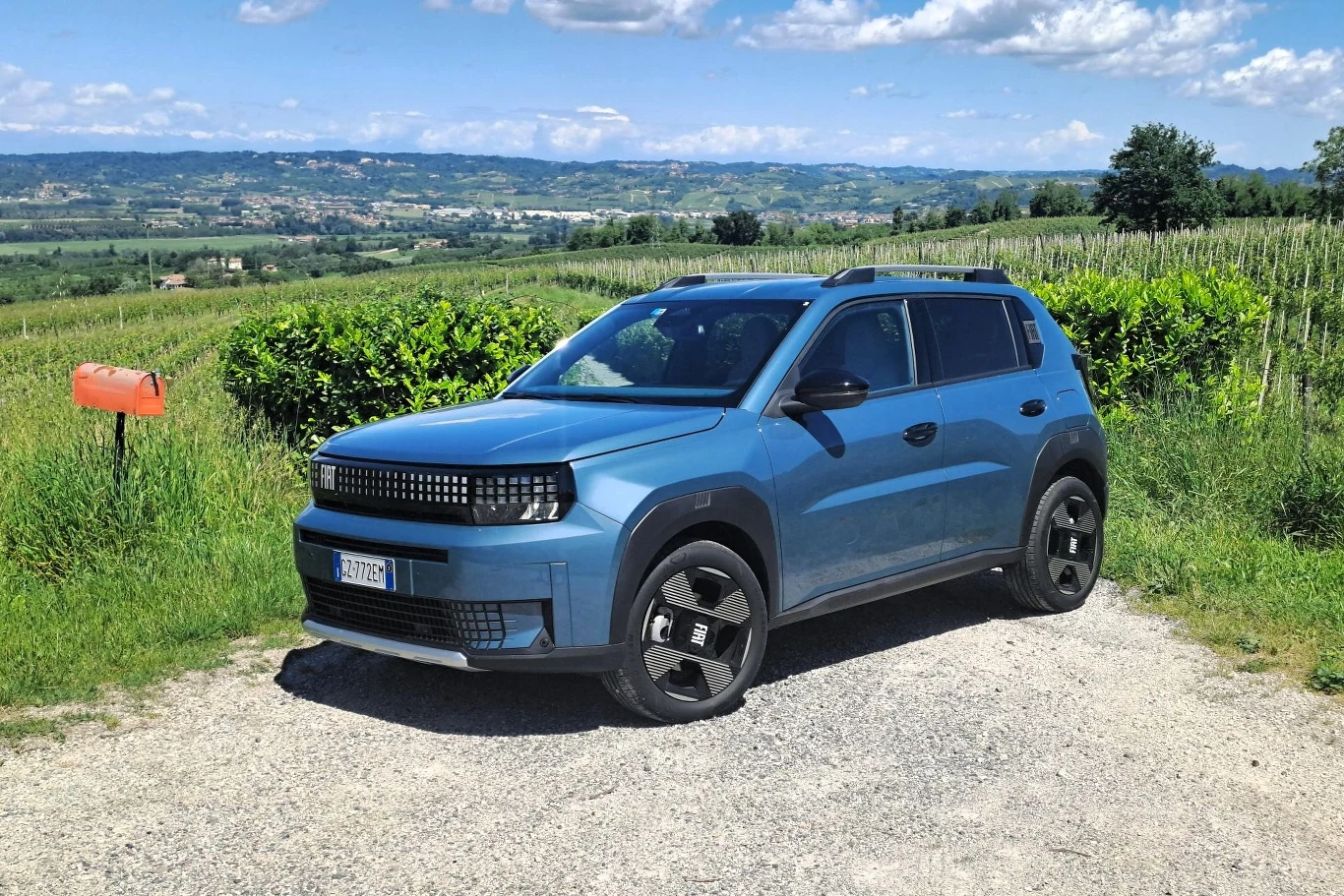 Niebieski nowoczesny samochód typu SUV zaparkowany na żwirowej drodze, w tle zielone pola, pagórkowaty krajobraz oraz niebieskie niebo z białymi chmurami, z lewej strony widoczna czerwona skrzynka pocztowa.