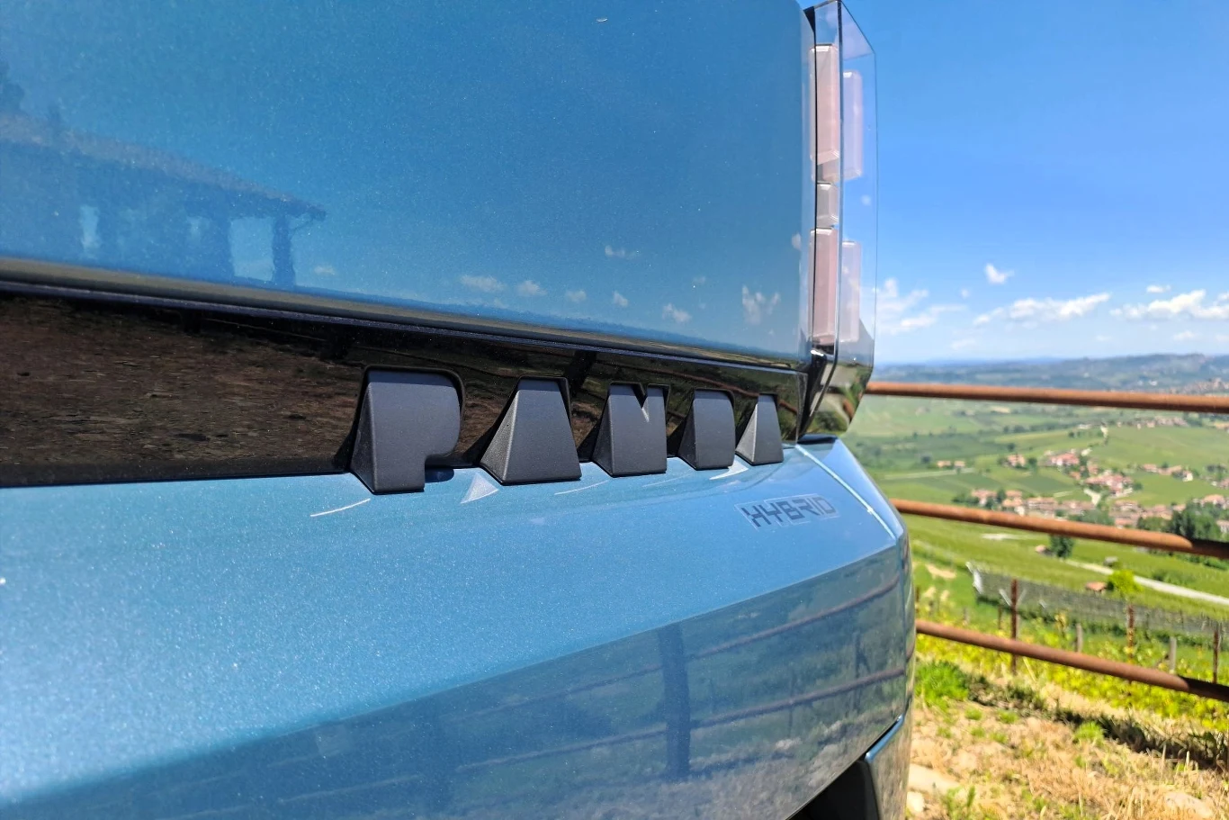 O tym, że mamy do czynienia z wersją hybrydową przypomni nam napis "HYBRID" na klapie bagażnika, tuż pod napisem "PANDA". Detal tylnej części niebieskiego samochodu Fiat Panda Hybrid ustawionego na tle wiejskiego krajobrazu z zielonymi łąkami i niebieskim niebem.