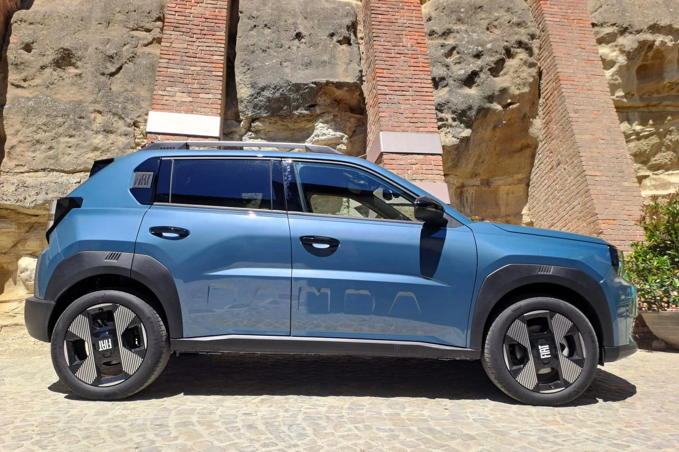Hybryda Grande Pandy o mocy systemowej 110 KM pozwala na rozpędzenie się do 100 km/h w 10 sekund. Niebieski samochód typu SUV zaparkowany na brukowanej drodze przy kamiennej ścianie z elementami czerwonej cegły, pojazd wyróżnia się nowoczesnym designem i dużymi kołami.