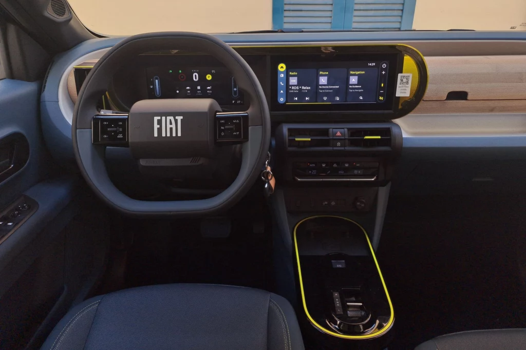 Ceny Fiata Grande Pandy startują od 79 900 zł. Kokpit nowoczesnego samochodu marki Fiat z cyfrowym wyświetlaczem, czarną kierownicą z logo producenta, dużym ekranem dotykowym oraz kontrastowymi, żółtymi akcentami w konsoli centralnej.