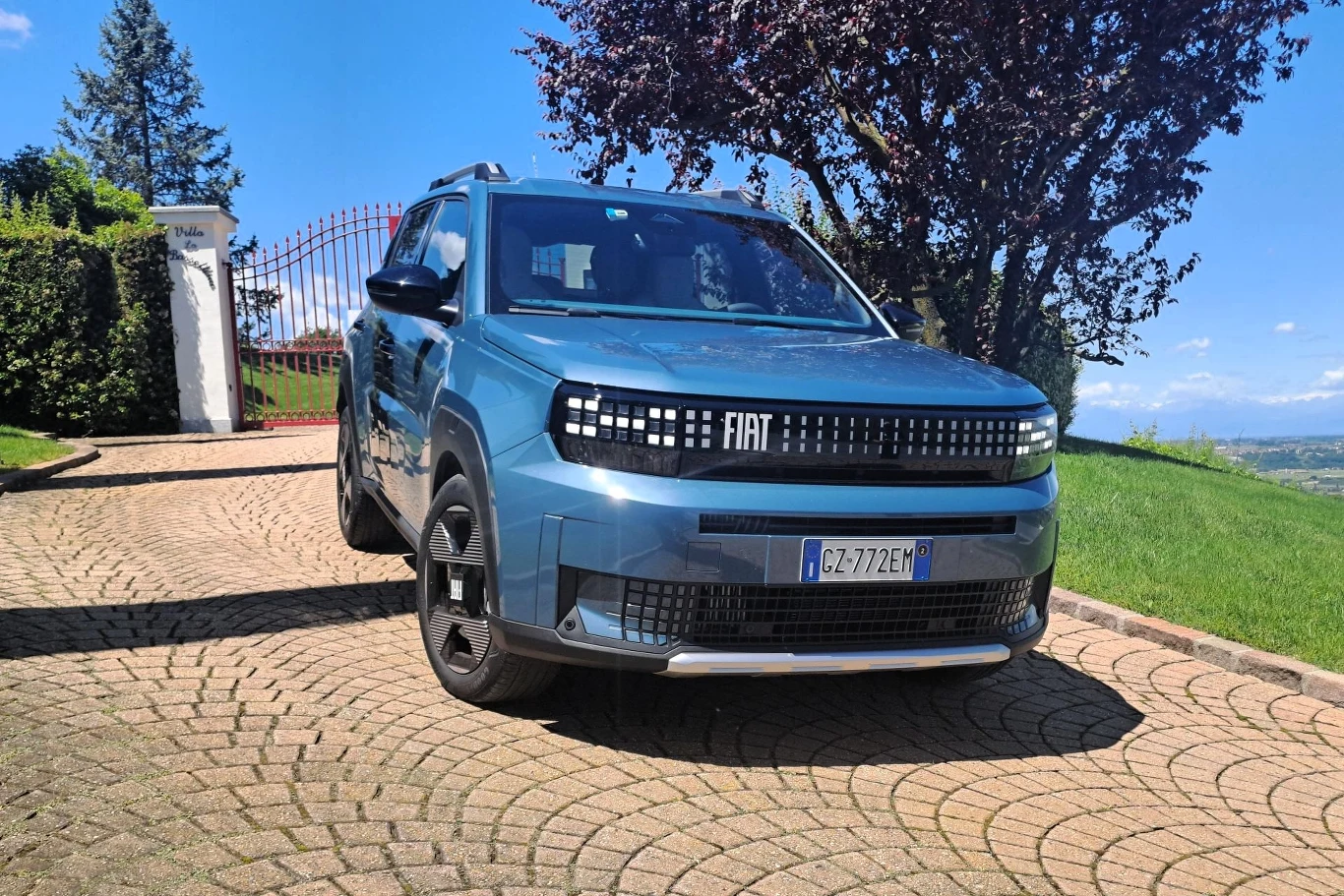 Fiat Grande Panda wykorzystuje ten sam układ hybrydowy co jego konstrukcyjny bliźniak, Citroen C3. Francuski model nie oferuje jednak tego napędu już w podstawowej wersji wyposażenia. Nowoczesny, niebieski samochód marki Fiat zaparkowany na brukowanym podjeździe przed zamkniętą bramą, w otoczeniu zieleni oraz drzew, z widoczną tablicą rejestracyjną.