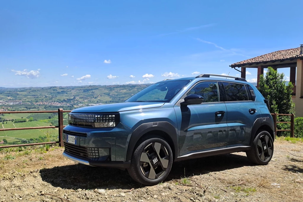 Fiat Grande Panda ma 3,99 metra długości. Dla porównania zwykła Panda ma 3,7 m. Nowoczesny miejski SUV w metalicznym niebieskim kolorze zaparkowany na tle zielonego krajobrazu z górzystym horyzontem i fragmentem stylowego domu po prawej stronie.