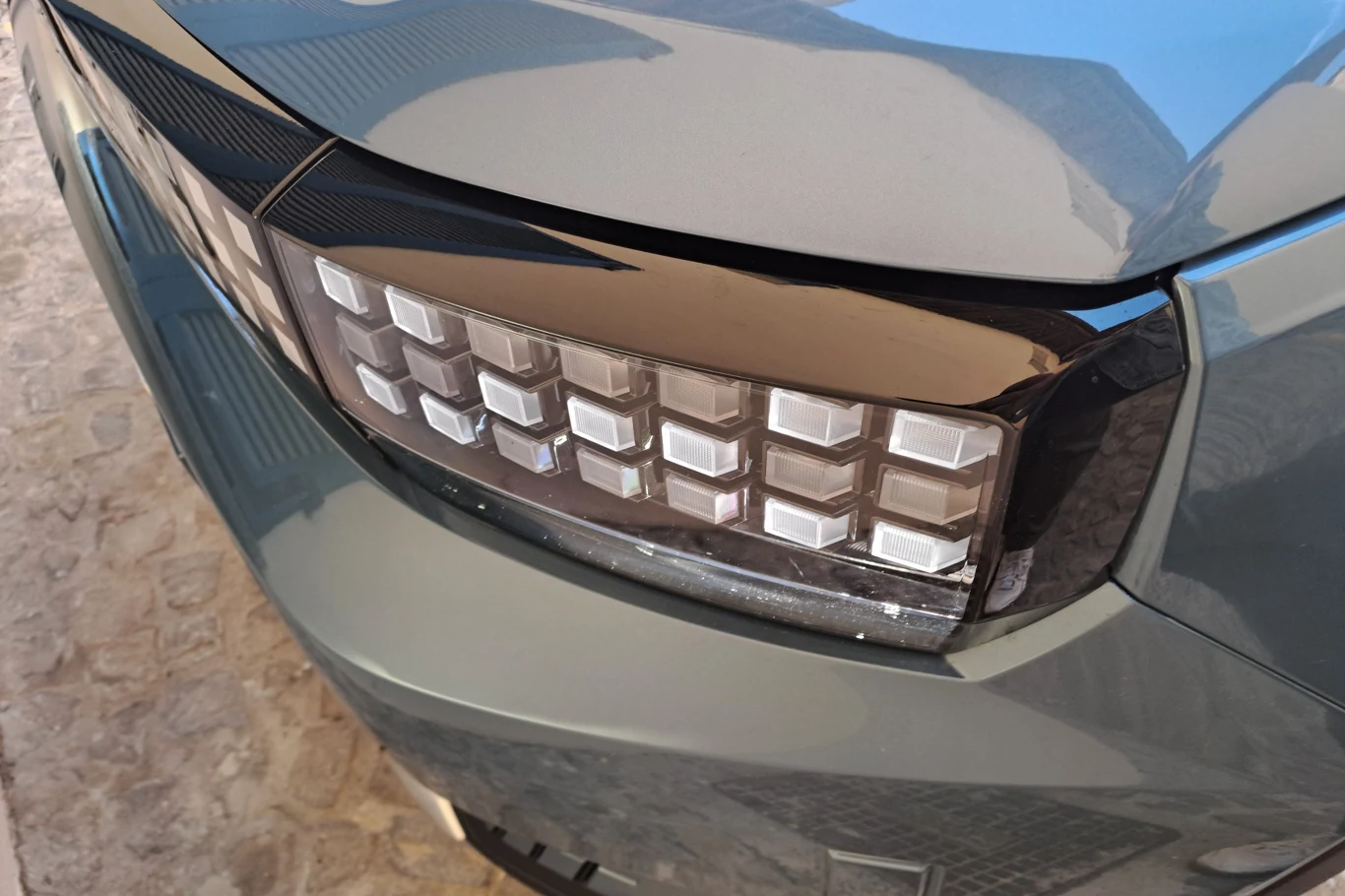 Pikselowe światła do jazdy dziennej, podobnie jak w wariancie elektrycznym, nawiązują do gier z lat 80. Nowoczesny reflektor samochodowy z wieloma prostokątnymi źródłami światła LED, umieszczony na gładkim, metalicznym nadkolu auta, otoczony odbiciami otoczenia.