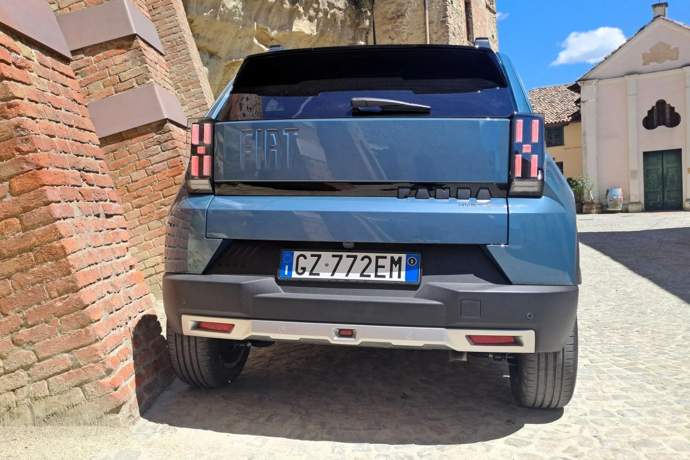 Średnie zużycie paliwa hybrydowej Grande Pandy wynosi, w zależności od wersji wyposażenia, od 5 do 5,1 litra na 100 km. Tył niebieskiego samochodu Fiat Panda z włoską tablicą rejestracyjną, zaparkowany na brukowanej uliczce przy murze, w tle fragmenty budynków i błękitne niebo.