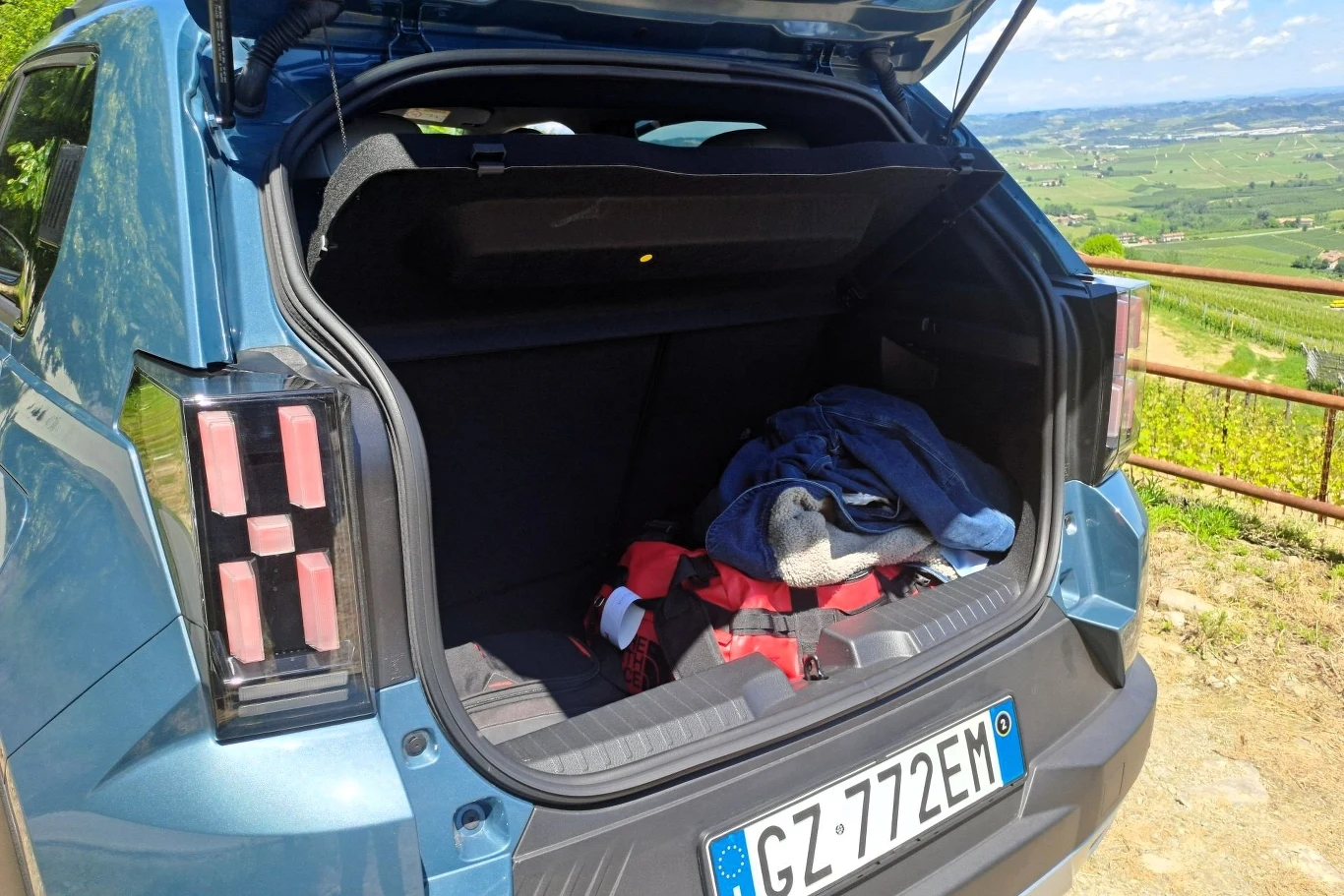 Bagażnik Fiata Grande Pandy Hybrid wynosi 412 litrów. W wariancie elektrycznym to 361 litrów. Otwarty bagażnik niebieskiego samochodu z włoską tablicą rejestracyjną, w środku rozłożone ubrania i torba, za pojazdem widoczny krajobraz z zielonymi polami i oddalonymi wzgórzami.