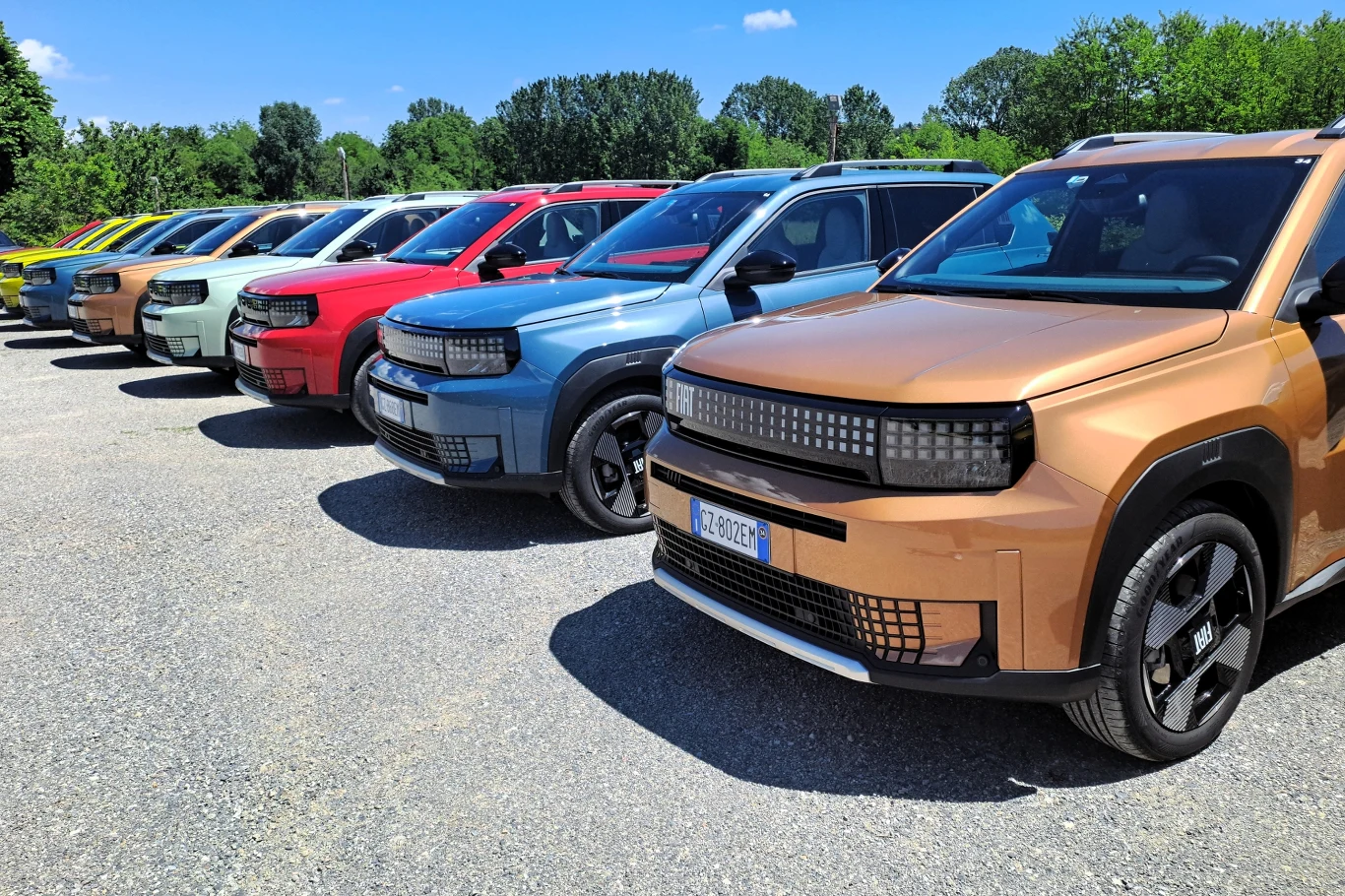 Nawet w topowej wersji wyposażenia ceny hybrydowego Fiata Grandy Pandy nie przekraczają 100 tys. zł. W takiej specyfikacji auto startuje od 96 900 zł. Rząd nowoczesnych samochodów terenowych w różnych kolorach ustawionych obok siebie na żwirowym parkingu, otoczony zielenią drzew i jasnym niebem.