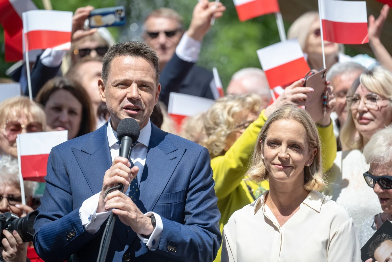 Rafał Trzaskowski na spotkaniu w Ciechocinku Mężczyzna w garniturze trzyma mikrofon i przemawia do zgromadzenia, obok niego stoi kobieta w jasnej koszuli, za nimi tłum ludzi z biało-czerwonymi flagami.