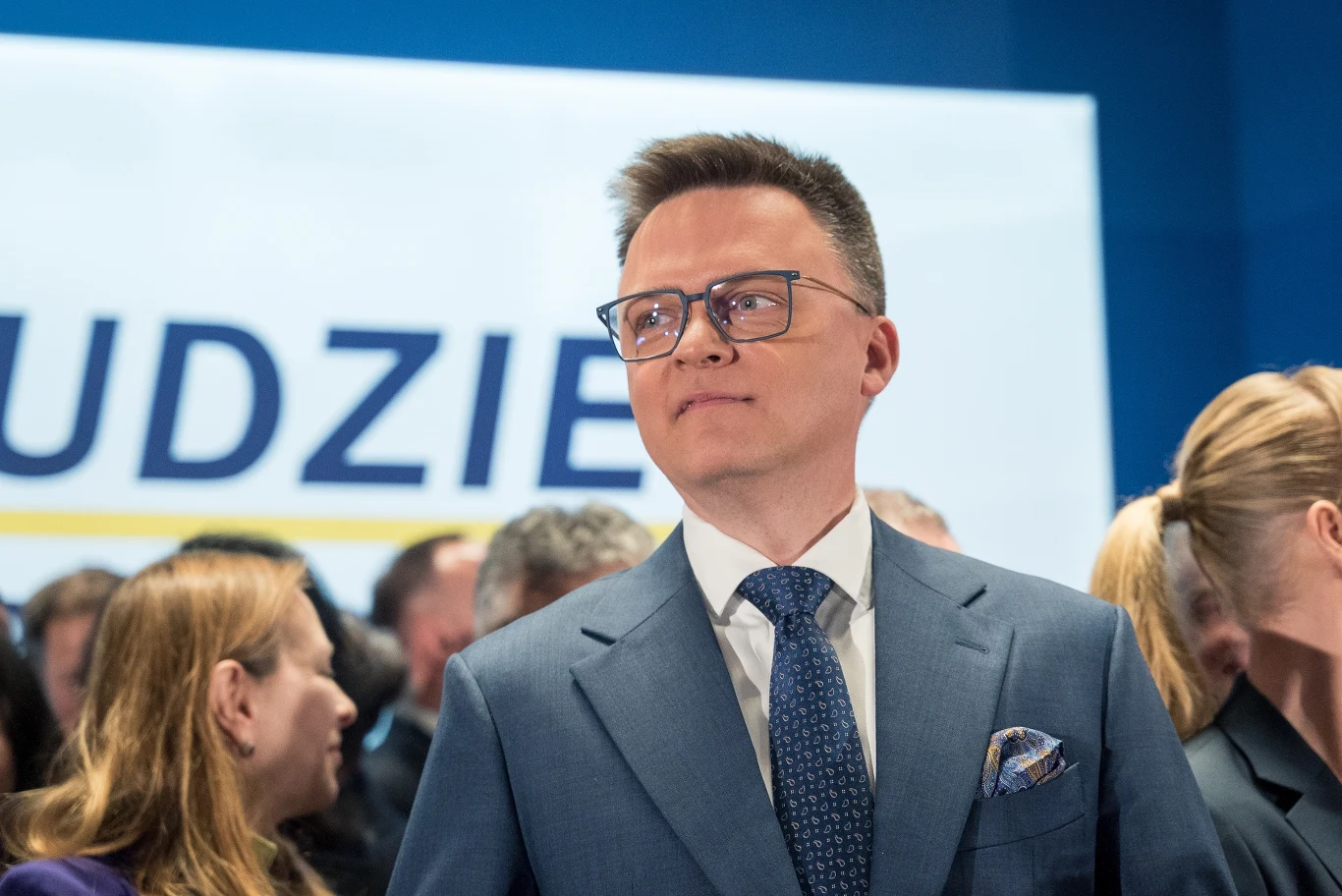Marszałek Sejmu Szymon Hołownia Mężczyzna w eleganckim garniturze i okularach stoi pośród grupy ludzi na tle napisu, sprawia wrażenie osoby przedsiębiorczej i pewnej siebie podczas oficjalnego wydarzenia.