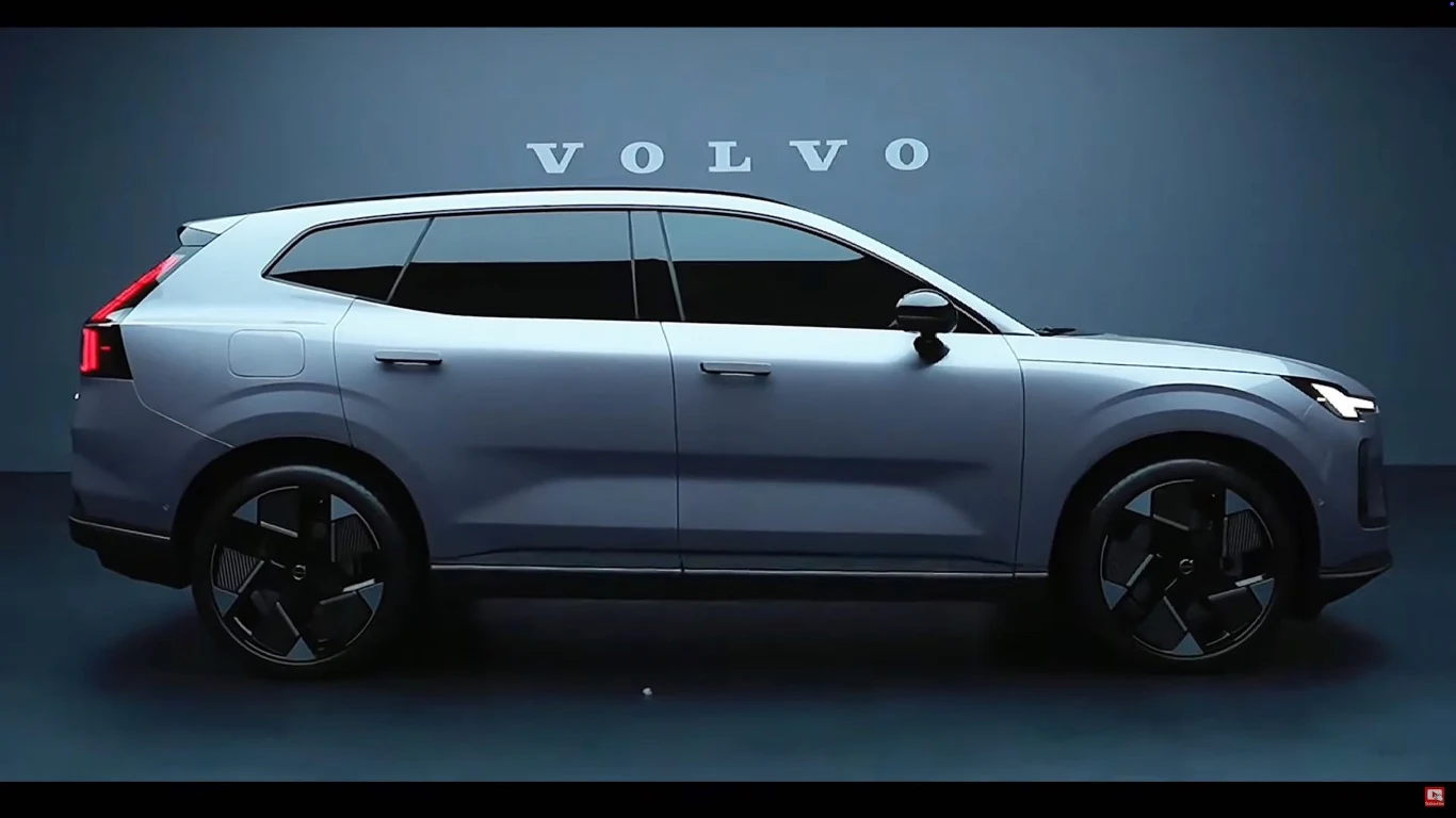 Nowe Volvo XC70 ma 481 cm, jest o 14 cm krótsze od XC90 Nowoczesny SUV marki Volvo w kolorze srebrnym, widoczny z profilu na tle niebieskiej ściany z dużym napisem VOLVO, charakterystyczne reflektory LED, masywne felgi oraz dynamiczna sylwetka pojazdu.