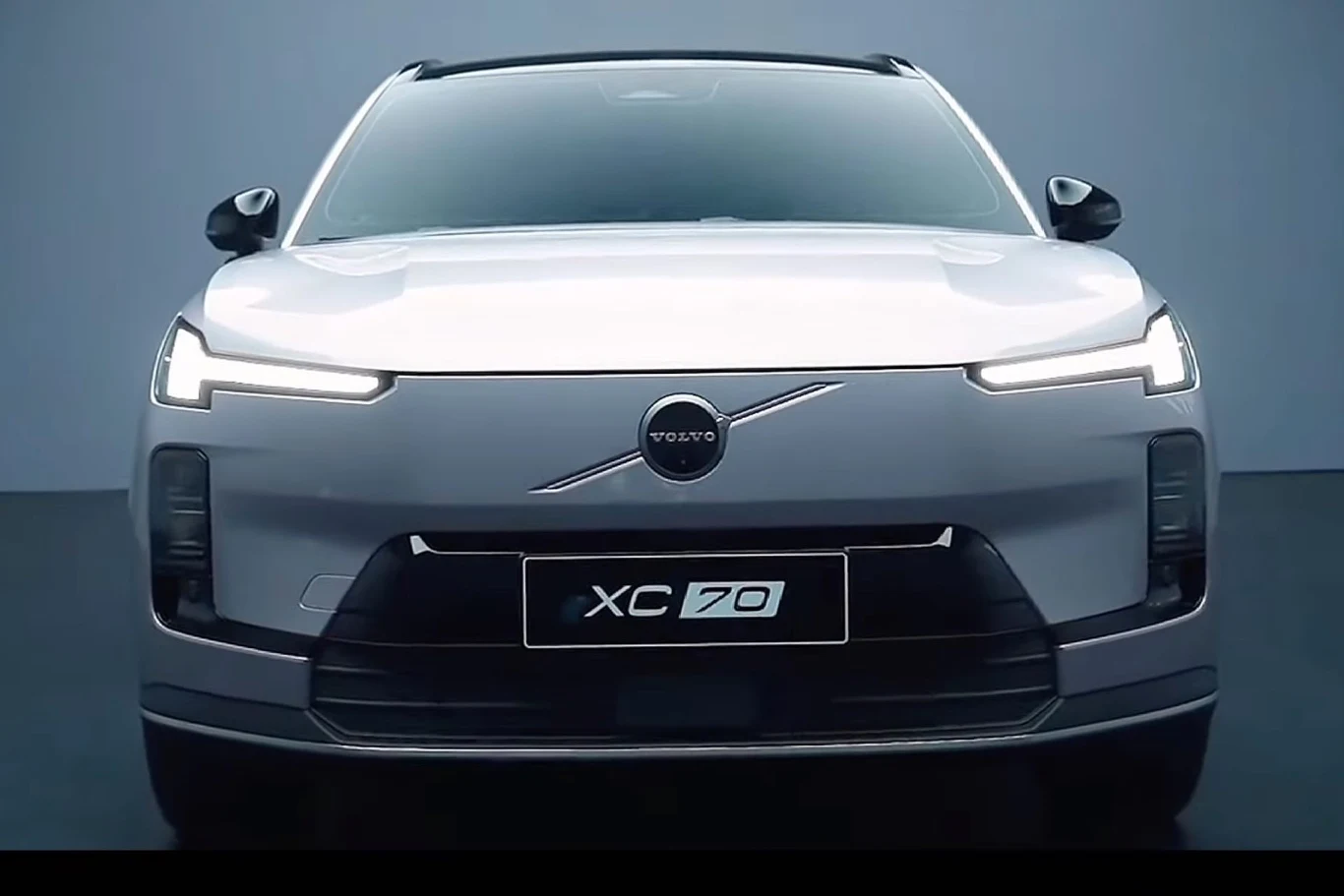 Stylizacja nadwozia XC70 nawiązuje do elektrycznych modeli marki Przód nowoczesnego, srebrnego samochodu Volvo XC70 z charakterystycznym logo marki i światłami LED, uchwycony na tle jednolitej, niebieskoszarej ściany.