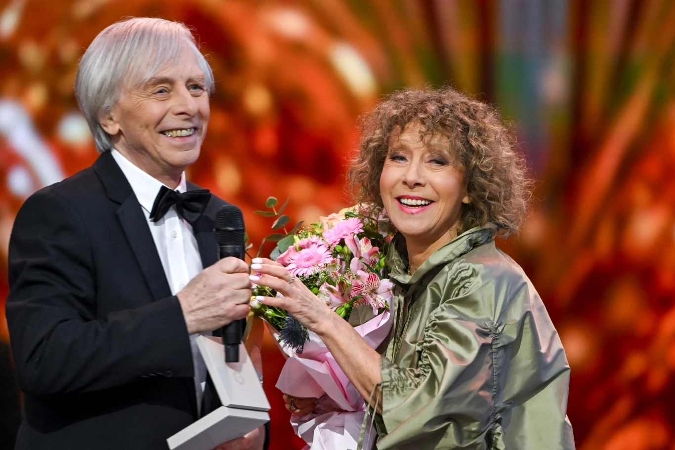 Włodzimierz Korcz i Alicja Majewska podczas Polsat Hit Festiwalu świętowali swoje 50-lecie na scenie Starszy mężczyzna w smokingu z mikrofonem oraz kobieta z bukietem kwiatów uśmiechają się, stojąc na scenie z kolorowym, rozmytym tłem.