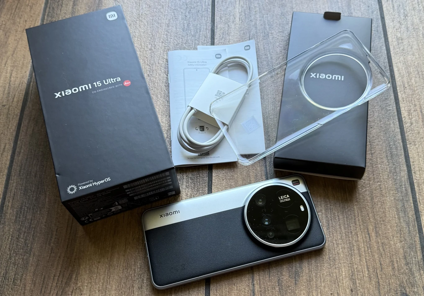 Zawartość opakowania z Xiaomi 15 Ultra. Smartfon Xiaomi 15 Ultra z dużym modułem aparatu Leica leży na drewnianej powierzchni obok akcesoriów: przezroczystego etui, kabla USB-C, instrukcji oraz pudełek.