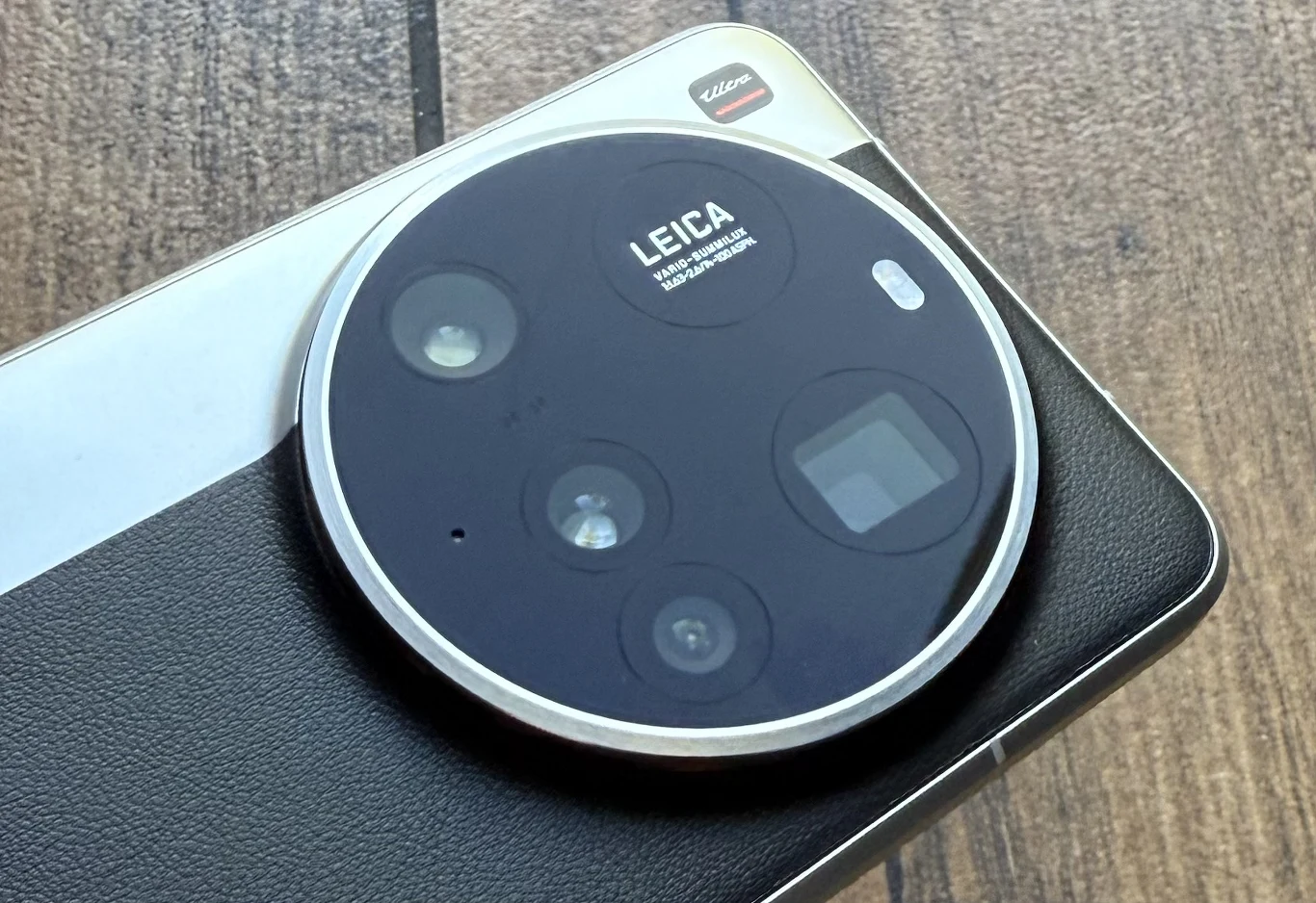 Xiaomi 15 Ultra to smartfon, który ma bardzo zaawansowany aparat o wielu możliwościach. Moduł aparatu fotograficznego Leica zamontowany w nowoczesnym smartfonie, wyraźnie widoczne kilka obiektywów i czujników optycznych na okrągłym panelu z wykończeniem imitującym skórę, smartfon leży na drewnianej powierzchni.