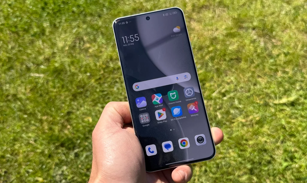 Ekran Xiaomi 15 Ultra jest bardzo jasny i pozwala na wygodnę pracę w pełnym nasłonecznieniu. Dłoń trzymająca nowoczesny smartfon z wyświetlaczem pokazującym ekran główny i aplikacje na tle zielonej trawy.