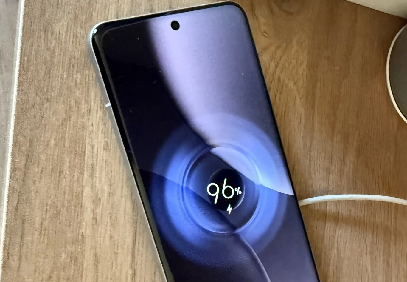 Xiaomi 15 Ultra ma dobrą baterię ze wsparciem dla szybkiego ładowania. Smartfon leżący na drewnianej powierzchni z widocznym ekranem pokazującym wskaźnik ładowania na poziomie 96%, do urządzenia podłączony jest biały przewód zasilający.