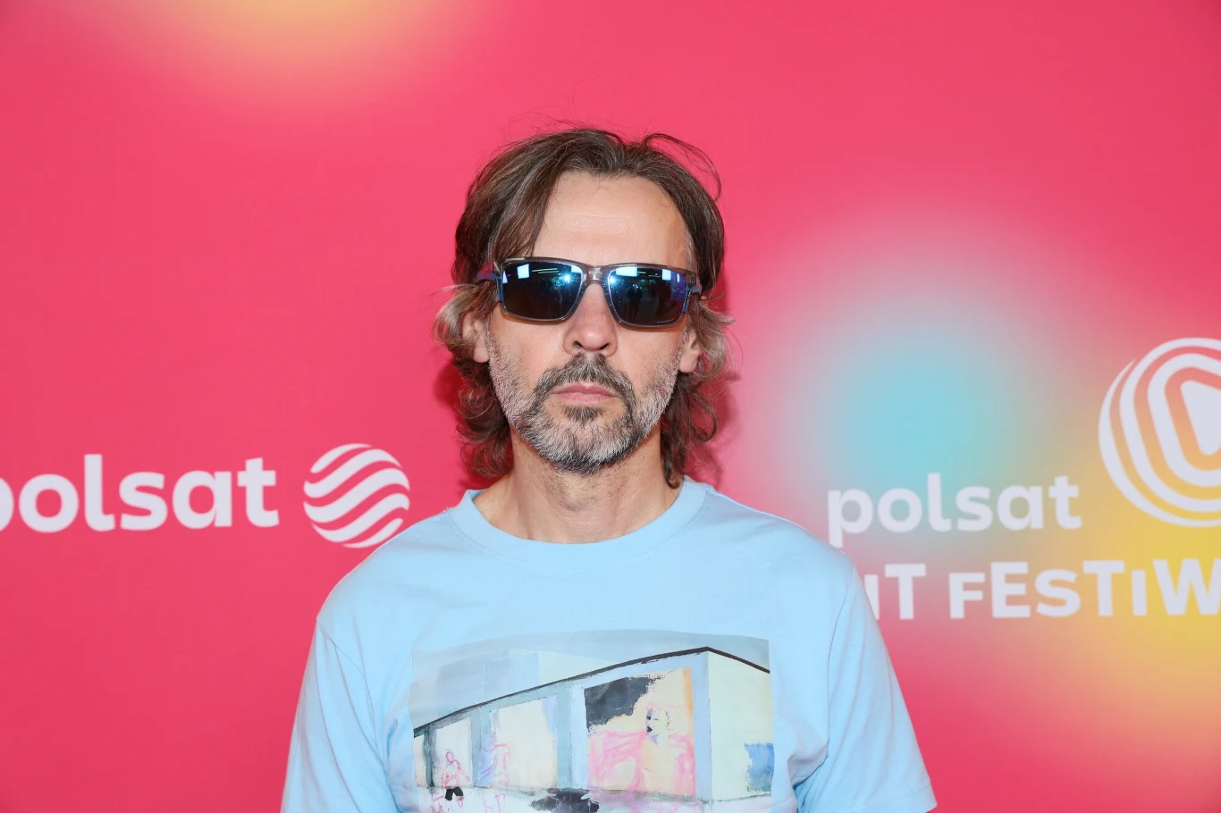Marcin Rozynek podczas Polsat Hit Festiwalu Mężczyzna z dłuższymi brązowymi włosami i brodą w ciemnych okularach przeciwsłonecznych pozuje na tle różowej ścianki z napisem polsat oraz FESTIW.
