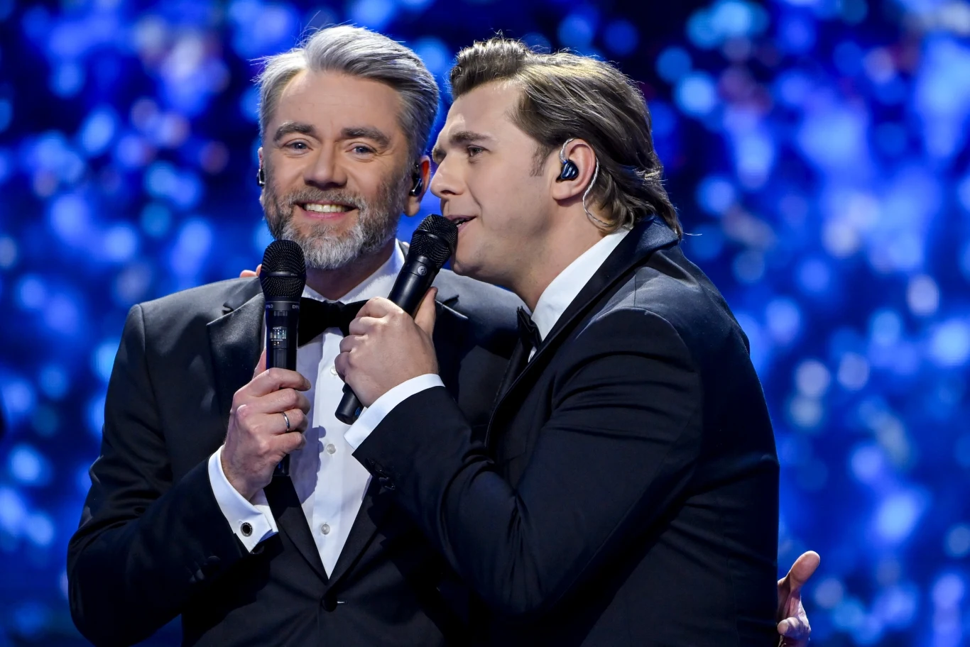 Kuba Badach i Andrzej Lampert podczas Polsat Hit Festiwalu Dwóch elegancko ubranych mężczyzn w garniturach śpiewa wspólnie do mikrofonów na tle rozświetlonej, niebieskiej sceny.