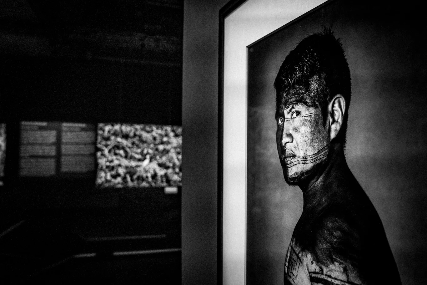 Portret mężczyzny z tatuażami na twarzy i ciele prezentowany na czarno-białej fotografii w ramie, wyeksponowanej w ciemnym pomieszczeniu wystawienniczym.