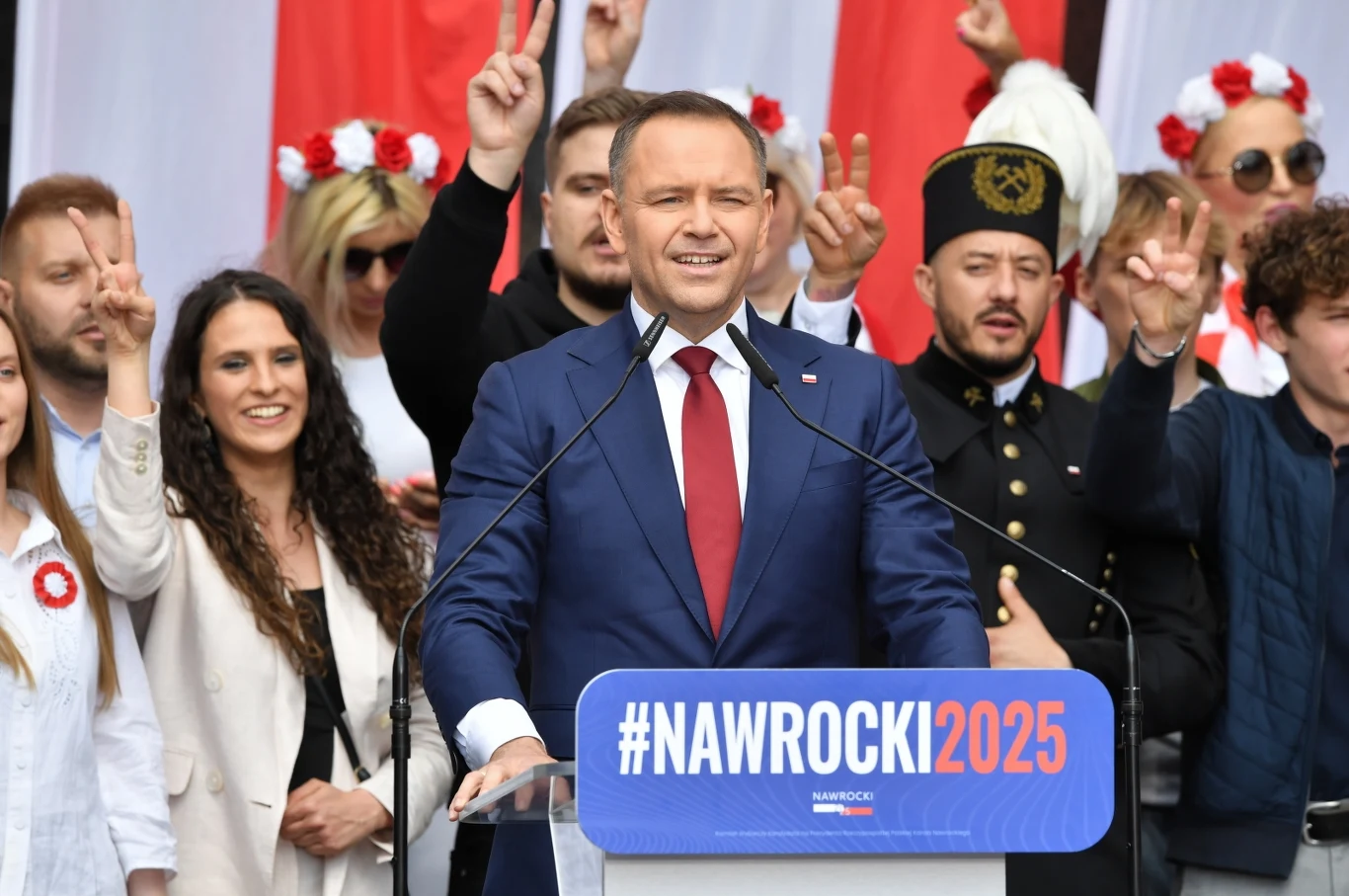 Karol Nawrocki przemówił do wyborców na placu Zamkowym Mężczyzna w garniturze przemawia przy mównicy z napisem #NAWROCKI2025, wokół niego stoi grupa uśmiechniętych osób, niektórzy wykonują gest zwycięstwa, w tle biało-czerwone dekoracje.