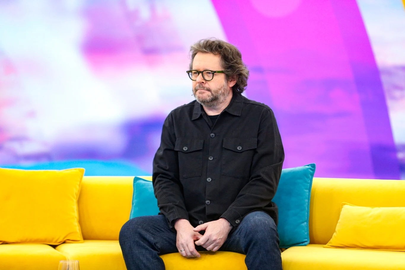 Grzegorz Turnau na żółtej kanapie w programie "halo tu polsat" Mężczyzna z okularami siedzący na żółtej sofie, w otoczeniu kolorowych poduszek, na tle fioletowo-niebieskiego, rozmytego ekranu.