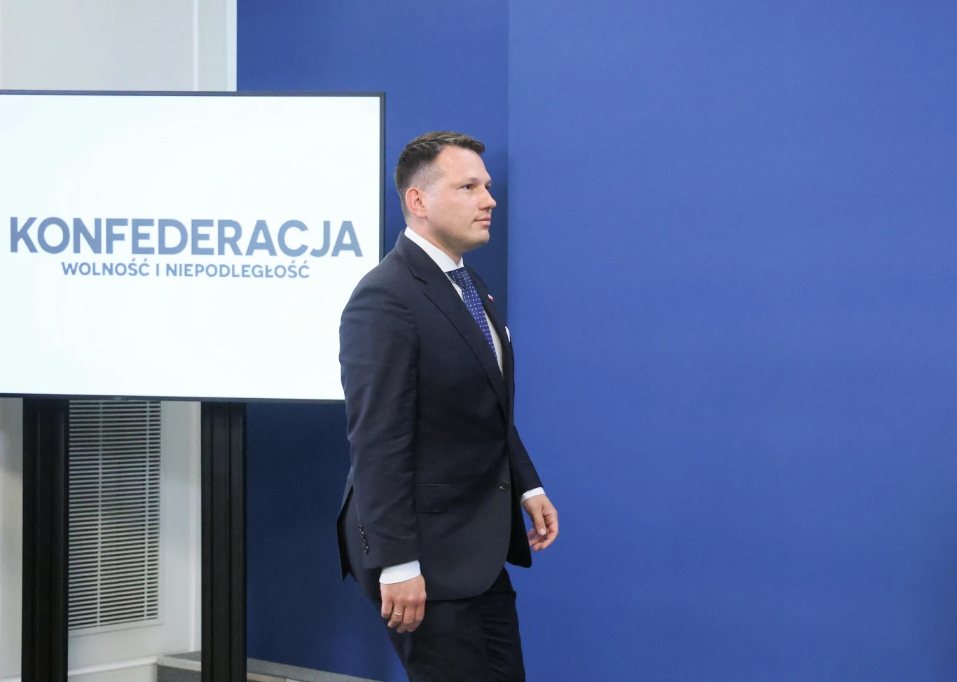 Wybory prezydenckie. Sławomir Mentzen przeprowadził rozmowy z kandydatami na urząd prezydenta RP Mężczyzna w garniturze idzie obok niebieskiej ściany, na tle znajdującego się ekranu z napisem 'KONFEDERACJA WOLNOŚĆ I NIEPODLEGŁOŚĆ'.