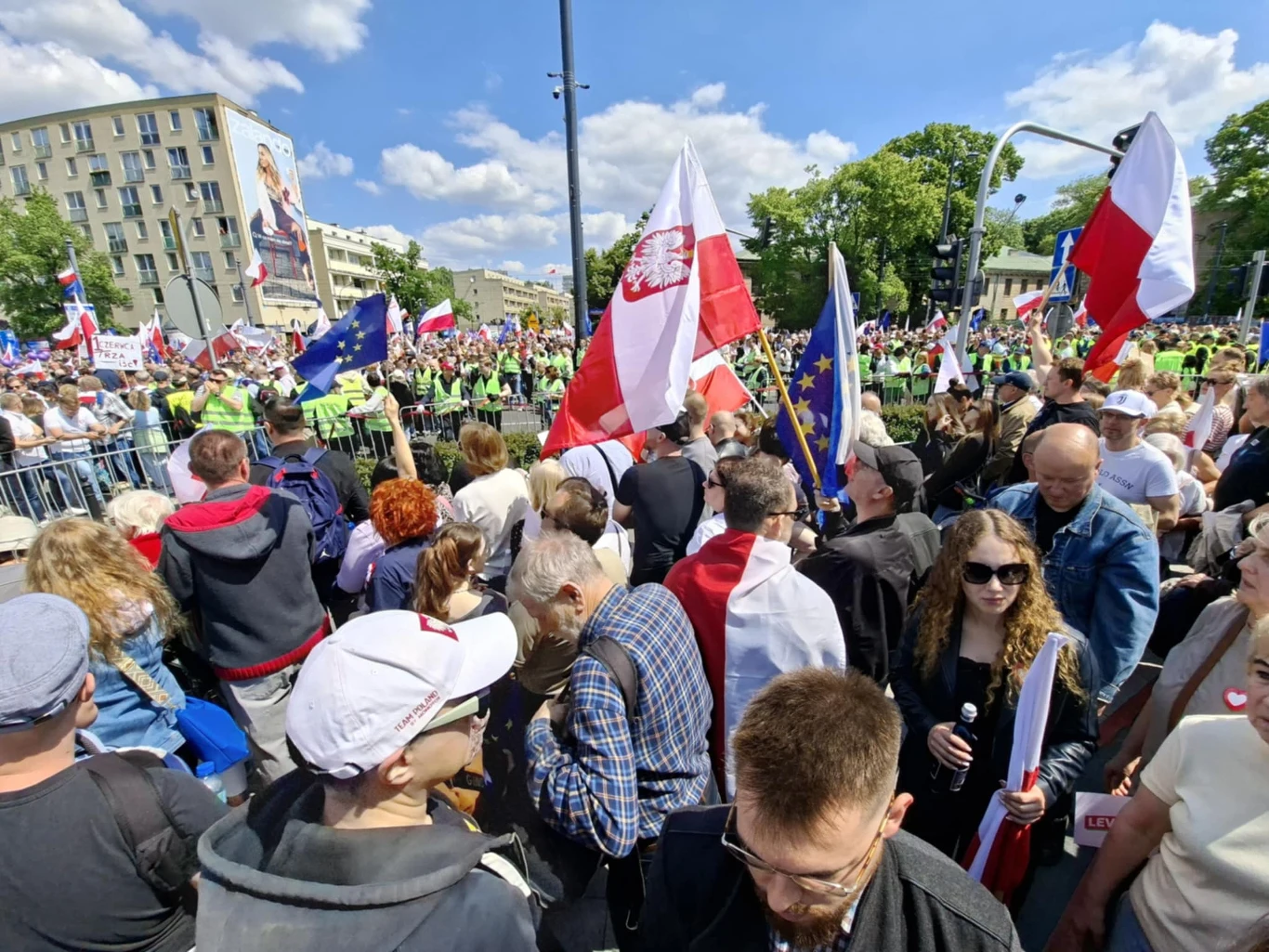 Tłum ludzi zgromadzony podczas demonstracji, wśród uczestników widoczne polskie oraz unijne flagi, w tle stoją policjanci tworzący kordon oraz budynki miejskie pod niebem z chmurami.