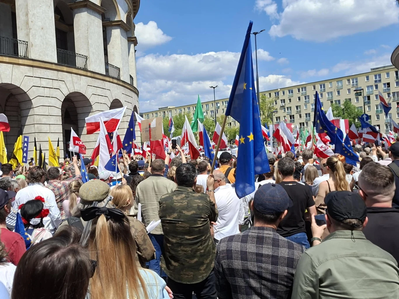 Tłum ludzi zgromadzonych przed budynkiem, wielu z nich trzyma flagi Polski, Unii Europejskiej oraz inne, charakterystyczne dla manifestacji i zgromadzeń publicznych; w tle widoczna zabudowa miejska.