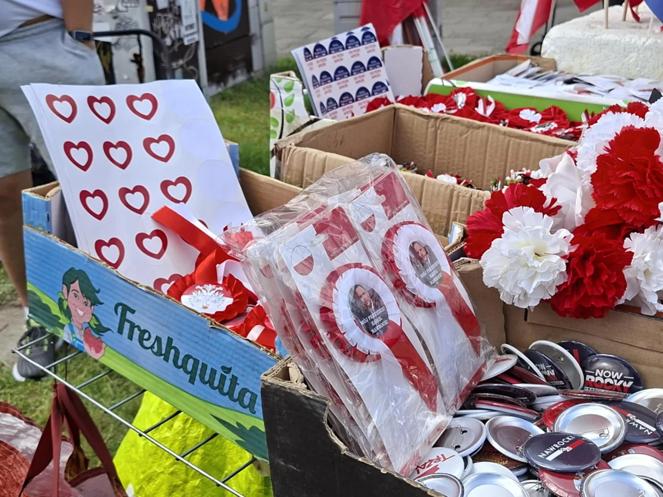 Kolorowe pudełka z przypinkami, sztucznymi kwiatami w barwach narodowych, plakatami z czerwonymi sercami i akcesoriami okolicznościowymi związanymi z wydarzeniem patriotycznym.