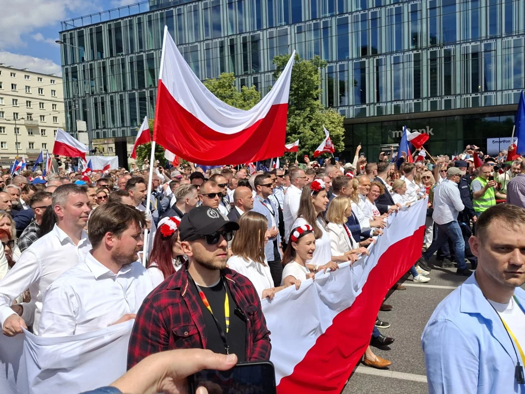 Czoło marszu Rafała Trzaskowskiego na ulicy Marszałkowskiej Tłum ludzi uczestniczący w patriotycznym marszu, trzymający dużą biało-czerwoną flagę oraz mniejsze polskie flagi, w tle nowoczesny szklany budynek i fragment klasycznej architektury, atmosfera radosna i podniosła.