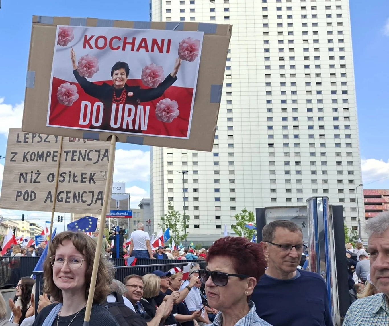 Tłum ludzi podczas demonstracji w centrum miasta, widoczne transparenty, w tym największy z napisem 'Kochani do urn' i wizerunkiem kobiety, w tle duży wieżowiec i flagi unijne oraz polskie.