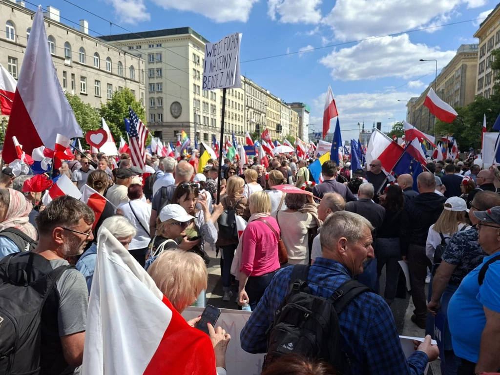 Zwolennicy Trzaskowskiego gromadzący się przed placem Konstytucji Tłum ludzi maszerujących ulicą miasta, otoczony licznymi flagami, głównie biało-czerwonymi oraz transparentami; w tle wysokie budynki i niebieskie niebo z obłokami.