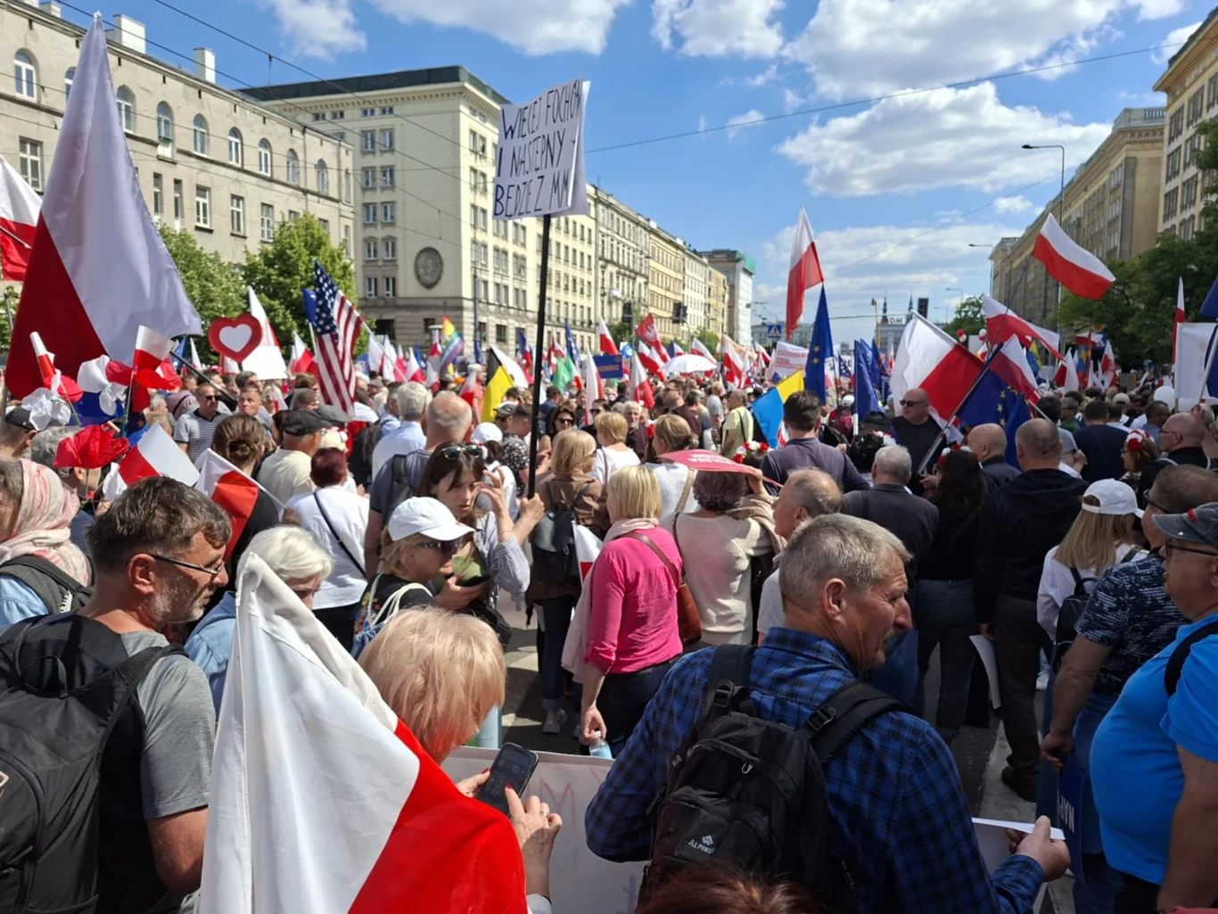Tłum ludzi maszerujących ulicą miasta, otoczony licznymi flagami, głównie biało-czerwonymi oraz transparentami; w tle wysokie budynki i niebieskie niebo z obłokami.