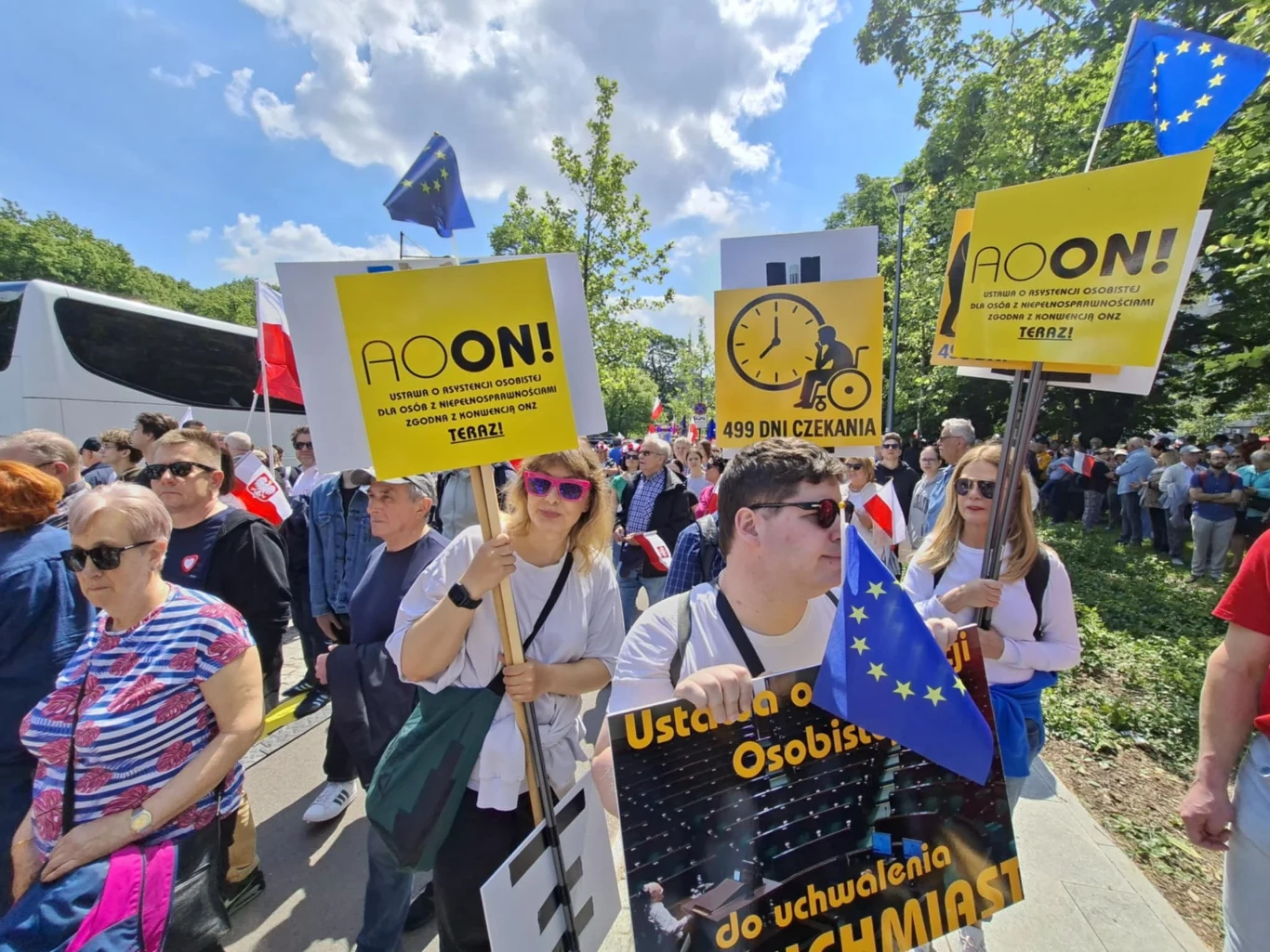 Tłum ludzi na ulicy podczas demonstracji, widoczne transparenty z żółtymi tablicami i napisy wzywające do zmian, flagi Unii Europejskiej, pogodny dzień, drzewa w tle, uczestnicy skupieni na marszu.