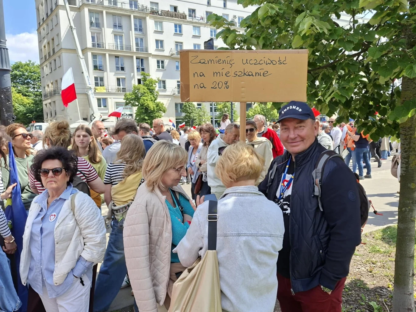 Tłum ludzi zgromadzony podczas protestu ulicznego, widoczna grupa kobiet i mężczyzn, w tle budynek mieszkalny i drzewa. Jeden z uczestników trzyma kartonowy transparent z hasłem o zamianie uczciwości na mieszkanie na 20%.