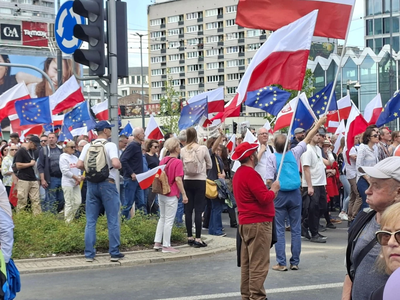 Tłum ludzi zgromadził się na ulicy, trzymając flagi Polski oraz Unii Europejskiej, w tle widoczne wysokie budynki oraz elementy miejskiej infrastruktury.